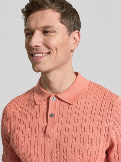 Männermodel trägt Slim Fit Poloshirt in Strick-Optik von Christian Berg Men, korallefarben, feine Strickstruktur, Nahaufnahme Oberkörper.