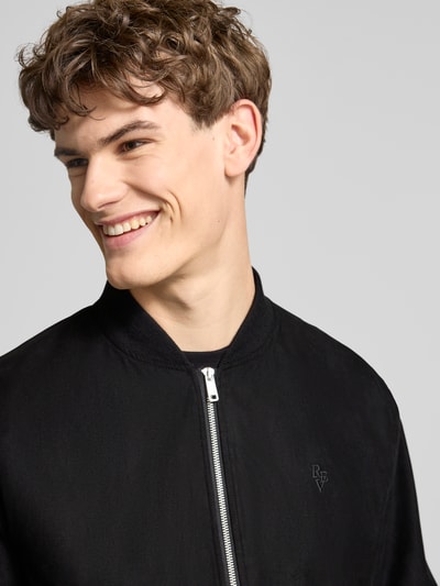 Männermodel trägt Bomberjacke mit Reißverschluss von REVIEW in schwarz, gerippte boorden, opstaande kraag, label-stiksel, detailopname bovenlichaam.