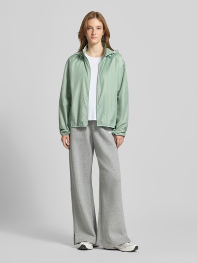 Frauenmodel trägt Regular Fit Jacke mit Stehkragen Modell 'Tempo' von NIKE TRAINING in Mintgrün, Kapuze mit Tunnelzug, Reißverschluss, Eingrifftaschen, elastische Abschlüsse, kombiniert mit weißem T-Shirt und grauer Hose, Rückenlänge 60 cm, Schulterbreite 54 cm, Brustweite 50 cm, Ärmellänge 59 cm, Ganzkörperansicht von vorne.