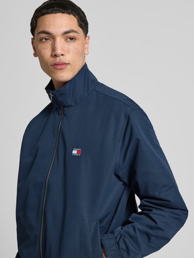 Męski model nosi Regular Fit Blouson mit Stehkragen od Tommy Jeans, jednokolorowy, z wpuszczanymi kieszeniami, naszywka z logo, stójka, długi rękaw, zapięcie zamek błyskawiczny, elastyczne wykończenie, efekt: jednolity kolor, ganzkörperansicht z przodu.