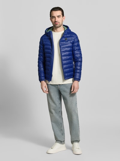 Männermodel trägt blaue Steppjacke mit Zweiwege-Reißverschluss Modell 'Charles' von Blauer Usa, Ganzkörperansicht von vorne, Regular Fit, Kapuze, Label-Detail, glatte und gesteppte Oberflächenstruktur, Rückenlänge 65 cm, Schulterbreite 47 cm, Brustweite 55 cm, Ärmellänge 65 cm.