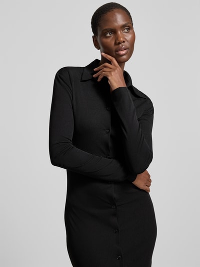 Frauenmodel trägt Hemdblusenkleid in Maxilänge von Nanushka, eng anliegende Passform, Hemdblusenkragen, durchgehende Knopfleiste, langärmelig, Farbe Schwarz, feine Textur, Nahaufnahme Oberkörper.