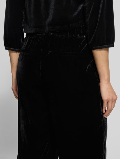 Kobiece model prezentuje Spodnie materiałowe z elastycznym pasem Model 'Mucia' od OPUS PANTS, czarne, wpuszczane i tylne kieszenie, luźny krój, tkanina o aksamitnej fakturze, długość wewnętrzna nogawki 79 cm, szerokość pasa 37 cm dla rozmiaru 36. Detailaufnahme z tyłu.