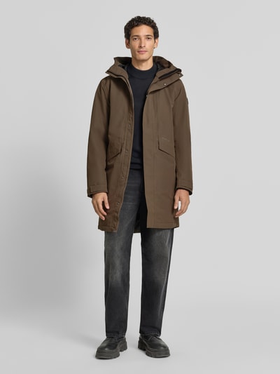 Männermodel trägt Regular Fit Parka mit Kapuze Modell 'Kenny' von Didriksons, braun, große Eingrifftaschen, Ganzkörperansicht von vorne. Rückenlänge 99 cm, Schulterbreite 48 cm, Brustweite 59 cm, Ärmellänge 72 cm.