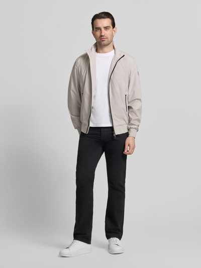 Männermodel trägt Regular Fit Jacke mit Stehkragen Modell 'Finlay' von SAVE THE DUCK in Beige, schwarze Hose, weißes Shirt, Ganzkörperansicht von vorne. Zweiwegereißverschluss, Reißverschlusstaschen, elastische Abschlüsse, sportlicher Look.