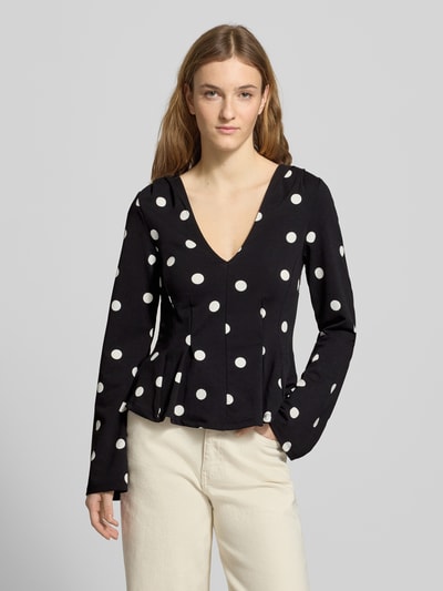 Modelka nosi Longsleeve mit V-Ausschnitt od Gina Tricot, czarny z białymi kropkami, materiał ze stretchem, krój taliowany, szeroki dekolt V, rozkloszowana część spódnicowa, długość tyłu w rozmiarze S: 53 cm, widok całej sylwetki z przodu.