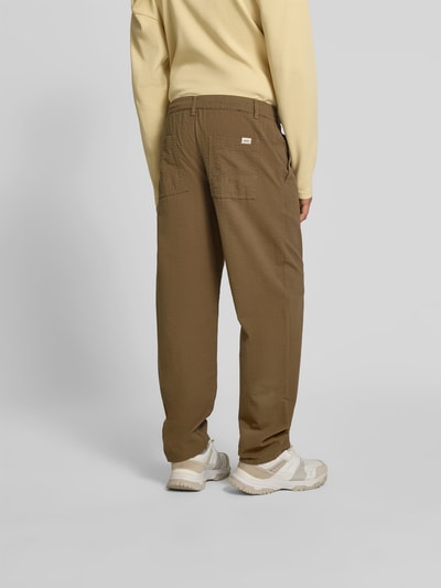 Männermodel trägt Stoffhose aus reiner Baumwolle von Forét in Braun mit Strukturmuster, Relaxed-Fit, Gesäßtaschen, Ganzkörperansicht Rückansicht.