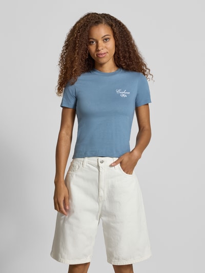 Frauenmodel trägt Slim Fit T-Shirt aus reiner Baumwolle Modell 'Gracious' von Carhartt Work In Progress, hellblau, Rundhalsausschnitt, Label-Print, kombiniert mit weißer kurzer Hose, Ganzkörperansicht von vorne, Rückenlänge bei Größe S: 53 cm.