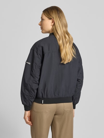 Frauenmodel trägt Jacke mit Pattentaschen Modell 'FACEE' von khujo in Schwarz, Regular-Fit, Stehkragen, Reißverschluss, Rückansicht Ganzkörperansicht. Rückenlänge bei Größe S 60 cm, Schulterbreite 50 cm, Brustweite 55 cm, Ärmellänge 56 cm, kombiniert mit Umhängetasche und sandfarbenen Hosen.