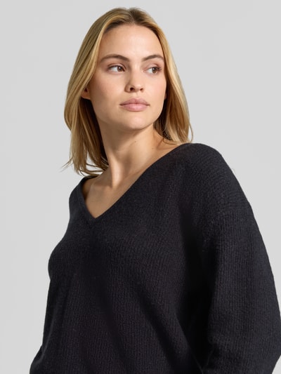 Modelka kobieca ma na sobie sweter z dzianiny z mieszanki materiałów z lnem, Efekt prążkowania, dekolt w serek, luźny krój, Model 'DAMSVILLE' od American Vintage, długość tyłu 61 cm, długość rękawów 54 cm, wykończenia elastyczne, widoczne kolory: czarny, tekstura miękka i prążkowana, zdjęcie w zbliżeniu na górną część ciała.