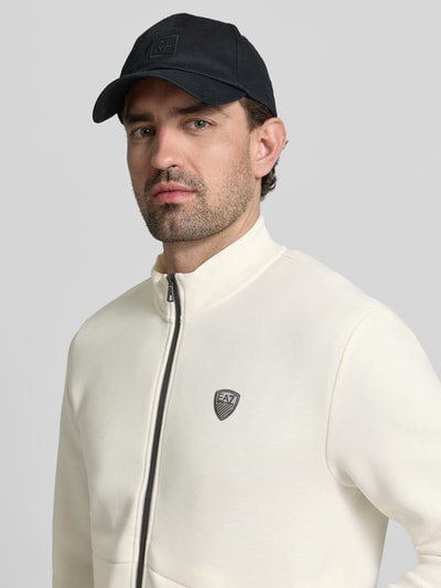 Männermodel trägt Sweatjacke mit Reißverschluss von EA7 Emporio Armani in Offwhite mit Stehkragen und Label-Detail, Nahaufnahme Oberkörper.