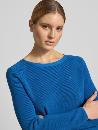 Vrouwenmodel draagt Regular Fit gebreide pullover uit puur katoen van Tommy Hilfiger, heldere blauwe tricotlook met logo-opschrift, lange mouw, ronde hals, fijn ribstructuur aan mouwboord en rompzoom, detailopname bovenlichaam.