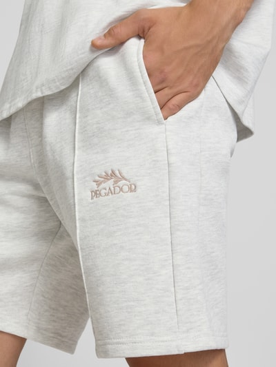 Männermodel trägt Sweatshorts mit Label-Stitching Modell 'Lido' von PEGADOR aus Baumwoll-Mix, Nahaufnahme Oberschenkel und Hand.