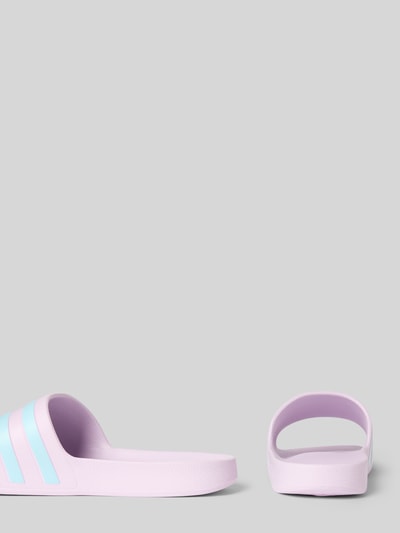 Produktfoto, Pantoletten mit Kontraststreifen von adidas Sportswear in Two-Tone-Optik, abgerundete Schuhspitze, flache Absatzform, Schlupf/offen, glatte Sohle, Materialverarbeitung Kunststoff, zarte Fliederfarbe mit hellblauen Streifen.