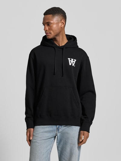 Männermodel trägt Hoodie mit Logo-Print und Känguru-Tasche von Wood Wood, schwarz mit weißem Print, lockere Passform, Ganzkörperansicht von vorne.