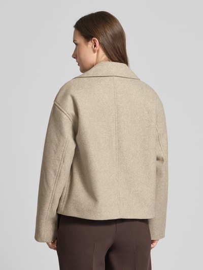 Frauenmodel trägt beigefarbenen Cropped Trenchcoat mit doppelreihiger Knopfleiste Modell 'VINCELINE' von Vero Moda, Boxy Fit, unifarben, Reverskragen, langarm, seitliche Eingrifftaschen, Ganzkörperansicht Rückansicht.