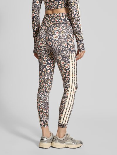 Frauenmodel trägt Slim Fit Sport-Leggings mit floralem Muster Modell 'FARM LEGGINGS' von adidas Sportswear, bunte Blumenfarben, Ganzkörperansicht Rückansicht.