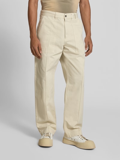 Männermodel trägt Straight Fit Hose aus reiner Baumwolle von Forét in Beige mit Eingrifftaschen, Gesäßtaschen, Knopf- und Reißverschluss, Ganzkörperansicht von vorne.