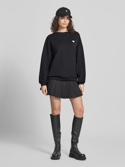 Frauenmodel trägt schwarzen Relaxed Fit Pullover mit Herz-Stickerei Modell 'TALIA' von Pieces, unifarben, Ganzkörperansicht von vorne.