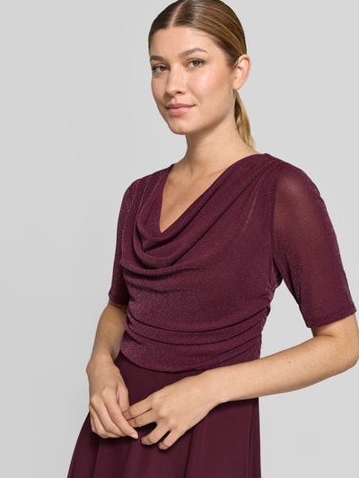 Kobieta nosi Fit and Flare Cocktailkleid mit Wasserfallausschnitt od Vera Mont w kolorze bordowym, połyskujący materiał, zbliżenie na górę.