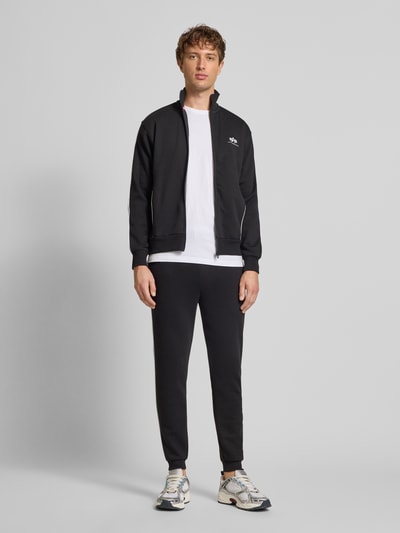 Männermodel trägt schwarze Sweatpants mit Logo von Alpha Industries, Regular-Fit, elastischer Bund, Tunnelzug, Ganzkörperansicht von vorne. Herren Sweatpants, Innenbeinlänge bei Größe M: 72 cm.