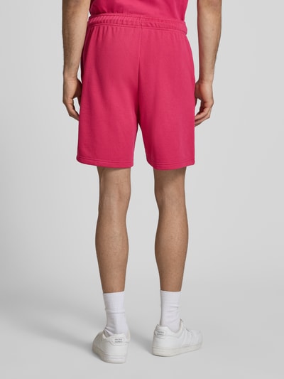 Mannenmodel draagt Sweatshorts mit Logo und elastischem Bund van CHAMPION in regular fit, roze katoenmix, elastische tailleband met trekkoord, logo detail, taillewijdte maat M 41 cm, binnenbeenlengte 20 cm, geheel van achteren bekeken.