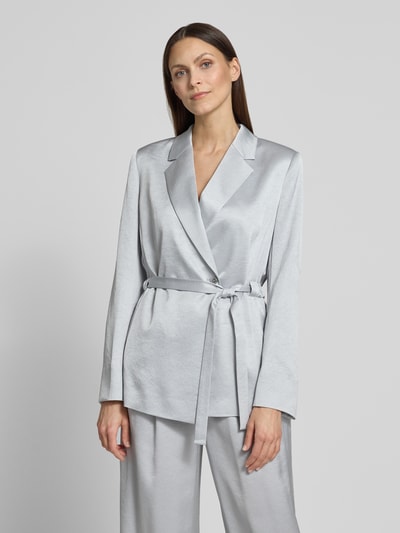Frauenmodel trägt silbergrauen Satin-Blazer mit Bindegürtel und passender Hose, glatte Oberfläche, Nahaufnahme Oberkörper.