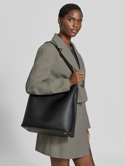 Frauenmodel trägt Handtasche in Leder-Optik Modell 'Nelma' von Seidenfelt, schwarz, breites Hauptfach, Logo-Detail, Ganzkörperansicht von vorne.