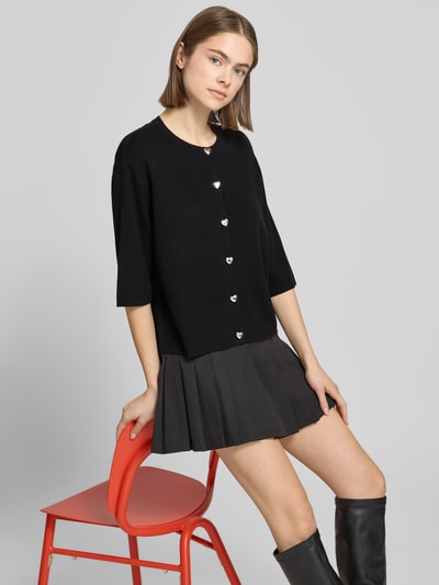 Modelka nosi Regular Fit Strickjacke aus Viskose-MIx Modell 'MAISIE' od ONLY, czarny drobny dzianina, guziki z ozdobami, krótki rękaw, dekolt okrągły, falisty dół i rękawy, ujęcie całej sylwetki z przodu.