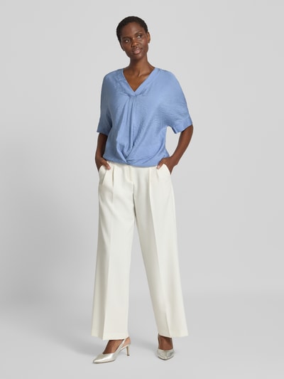 Frauenmodel trägt Blusenshirt mit V-Ausschnitt von Christian Berg Woman in Hellblau, kombiniert mit weißer Hose; fließende Textur, Ganzkörperansicht von vorne. Kontext: Damen Blusenshirt, Regular Fit, 1/2-Arm, Drapierung, Rückenlänge 31 cm, Ärmellänge 61 cm.