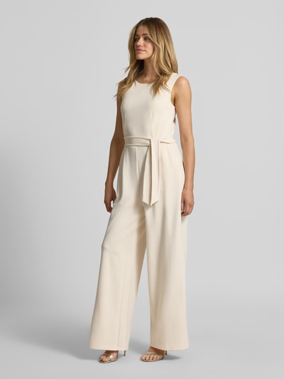 Modelka nosi Jumpsuit aus Viskose-Mix mit Bindegürtel von Jake*s Collection, mieszanka wiskozy z dodatkiem streczu, krój regular fit, okrągły dekolt, wiązany pasek, długość tyłu w rozmiarze 36: 40 cm, szerokość w rozmiarze 36: 30 cm, całkowita długość w rozmiarze 36: 150 cm, długość wewnętrzna nogawki w rozmiarze 36: 80 cm, kremowy kolor, szerokie nogawki, gładka tekstura, pełny widok z przodu.
