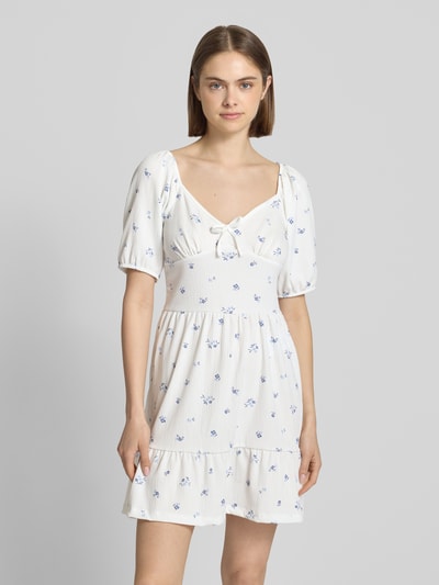 Frauenmodel trägt Minikleid mit Puffärmeln Modell 'HILLARY' von Only in Weiß mit floralem Allover-Muster und Schleifenapplikation, Crêpe-Textur, Ganzkörperansicht von vorne.