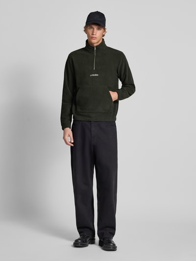 Męski model nosi Regular Fit Troyer mit Känguru-Tasche Modell 'SOHO HALFZIP FLEECE' od Jack & Jones w kolorze czarnym, długi rękaw, zamek, kieszeń typu kangurka, label-stitching, długość tyłu 67 cm, długość rękawa 67 cm, widok całej sylwetki z przodu.