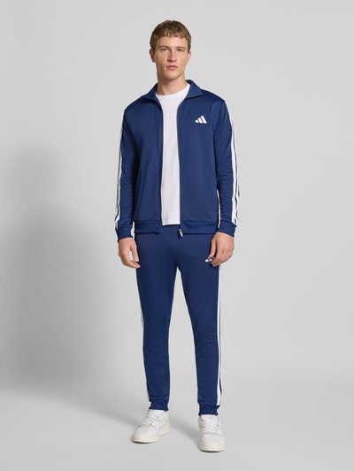Męski model prezentuje 'Trainingsanzug mit elastischen Abschlüssen' od adidas Sportswear, prosty krój, granatowy, całkowity widok z przodu