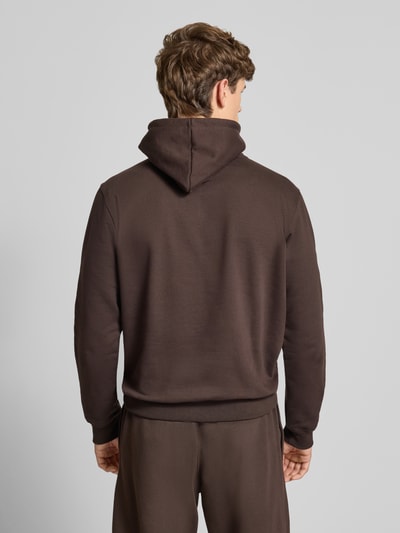 Męski model nosi Hoodie mit Kapuze od adidas Originals z mieszanki bawełny, luźny krój, ciemnobrązowy kolor, kangurkowa kieszeń, ściągacze, widok całej sylwetki z tyłu.
