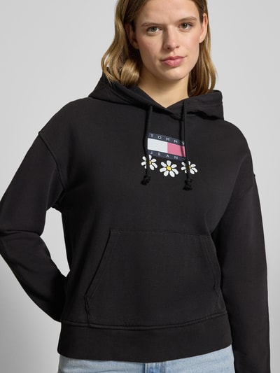 Modelka kobieca nosi Hoodie mit Label- und Motiv-Print Modell 'DAISY FLAG' od Tommy Jeans, czarny, luźny fason, bawełna ze stretchem, kaptur, nadruk, kieszeń kangurka, żebrowane wykończenia; ujęcie z przodu, zbliżenie na górną część ciała.