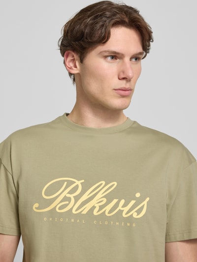 Męski model nosi T-Shirt mit Label-Print und Rundhalsausschnitt von BLKVIS z bawełny, krój regular fit, beżowy, nadruk z logo, zbliżenie na górną część ciała. Długość tyłu w rozmiarze M: 68 cm.