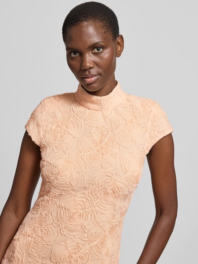 Frauenmodel trägt Maxikleid mit floralen Allover-Details von ROTATE im elastischen Material-Mix, taillierte Passform, Stehkragen, kurzärmelig, leicht strukturierte Blüten-Textur, Farbe Apricot, Nahaufnahme Oberkörper.
