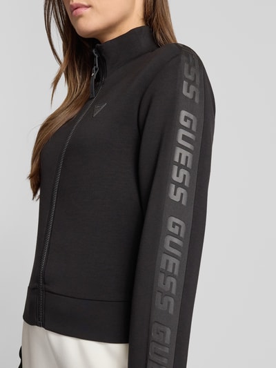 Frauenmodel trägt Regular Fit Sweatjacke aus Viskose-Mix von Guess, Schwarz mit Reißverschluss und Logo-Applikation, Nahaufnahme Oberkörper.