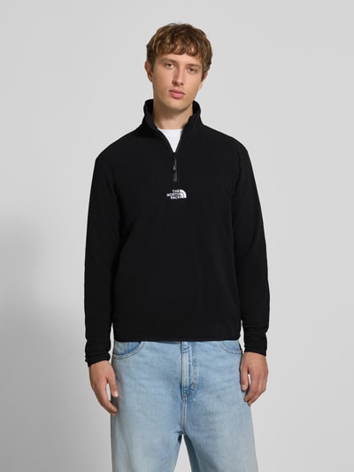 Männermodel trägt Fleecepullover mit Label-Stitching von The North Face in Schwarz, Stehkragen, Reißverschluss, Ganzkörperansicht von vorne