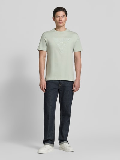Männermodel trägt T-Shirt mit Logo und Rundhalsausschnitt von Guess aus reiner Baumwolle, Regular-Fit, Rückenlänge 70 cm, Ganzkörperansicht von vorne.