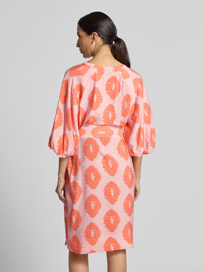 Frauenmodel trägt Knielanges Kleid aus Viskose-Mix von Christian Berg Woman in Koralle und Rosa mit Taillenband, taillierte Passform, Ganzkörperansicht Rückansicht, Serafino-Ausschnitt, halblange Ärmel, strukturierte Textur.