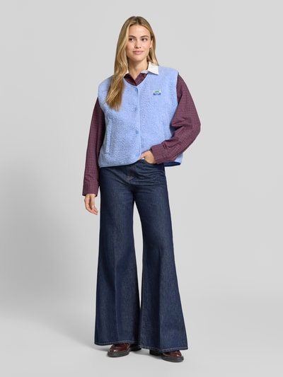 Frauenmodel trägt Relaxed Fit Weste aus Teddyfell Modell 'Hoktown' von American Vintage in Hellblau über Hemd und Jeans, Ganzkörperansicht von vorne. Damen Weste mit Logo-Patch, Knopfleiste sowie weicher Teddyfell-Textur, Rückenlänge 56 cm, Schulterbreite 43 cm, Brustweite 63 cm.