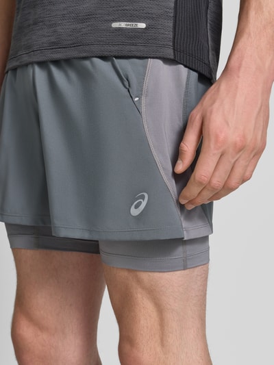 Męski model nosi Regular Fit Shorts mit integrierten Tights Model 'Road' von ASICS TIGER, krój regular fit, elastyczny pas, detal z logo, Szerokość w rozmiarze M: 38 cm, Długość wewnętrzna nogawki: 12 cm, odcień szarości, gładka struktura, z przodu, zbliżenie na górną część uda.