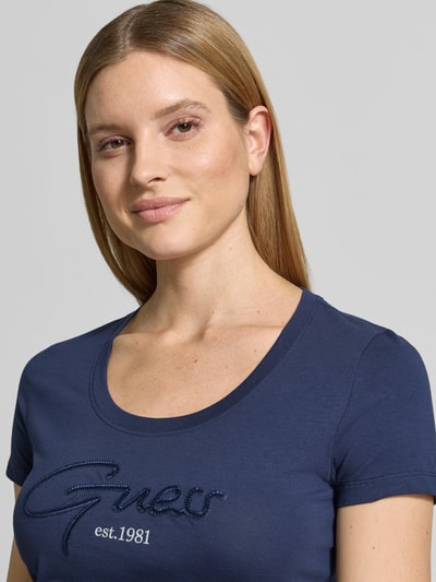 Vrouwenmodel draagt T-shirt met U-hals model 'SCRIPT' van Guess, mix van katoen en elastaan, nauwsluitend model, labelstitching, kleur blauw, dames t-shirt, detailopname bovenlichaam.