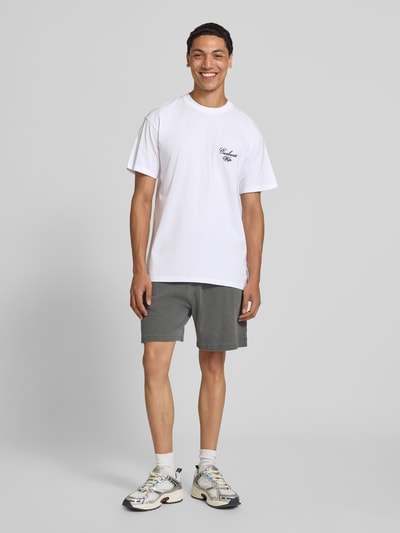 Männermodel trägt T-Shirt mit Logo-Stitching Modell 'Gracious' von Carhartt Work In Progress aus weißer Baumwolle, Ganzkörperansicht von vorne.