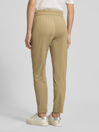 Modelka nosi Slim Fit Stoffhose mit Reißverschlusstaschen von Marc Cain, elastyczna mieszanka materiałów, krój slim fit, elastyczny pas, tunel, detale z logo, kieszenie na suwak, szerokość w rozmiarze 36: 37 cm, długość nogawki od wewnątrz w rozmiarze 36: 68 cm, beżowy kolor, gładka tekstura, widok całej sylwetki tyłem.