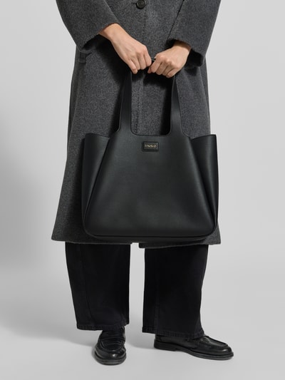 Frauenmodel trägt Shopper mit Logo-Detail von HUGO in schwarzer Lederoptik mit Tragehenkeln, Hauptfach und Innenfach, Ganzkörperansicht von vorne.