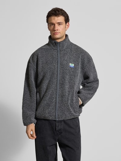 Männermodel trägt Regular Fit Jacke mit Logo-Patch von American Vintage in Dunkelgrau, Kunstfelltextur, Stehkragen, Ganzkörperansicht von vorne. Rückenlänge 69 cm, Schulterbreite 42 cm, Brustweite 67 cm, Ärmellänge 72 cm.