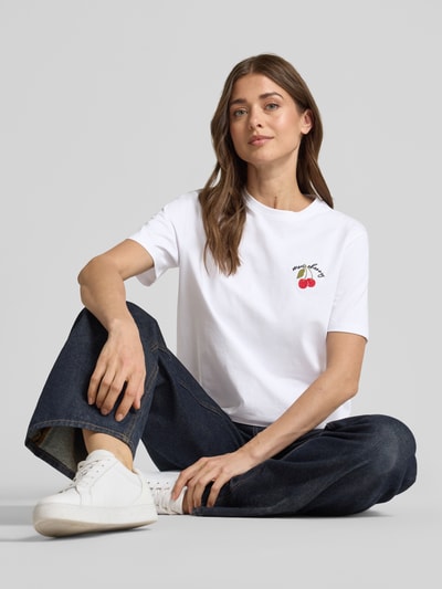 Vrouwenmodel draagt T-shirt met Stitching model 'NORA' van Vila, mix van katoen en elastaan, regular fit, ronde hals, ruglengte bij maat S 58 cm, kleur wit met kersenprint, gladde textuur. Zicht van voren, detailopname bovenlichaam.